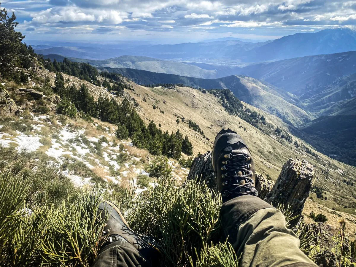 Les pieds du chasseur posés au bord d'une montagne, avec une vue imprenable sur la vallée et les sommets lointains sous un ciel vaste.