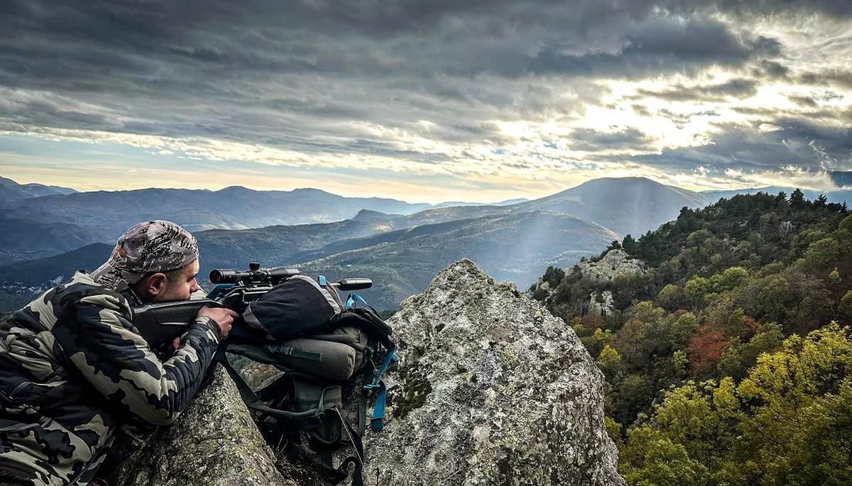Un individu camouflé est accroupi sur un point de vue rocheux, scrutant à travers une paire de jumelles un vaste paysage montagneux sous un ciel nuageux spectaculaire.